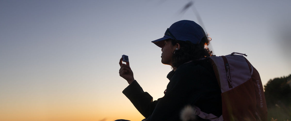 A photograph of a young women in her 20's holding a Mighty 3 in the air. She's wearing a Patagonia backpack, Oakley sunglasses are propped above her purple cap. The background sunset lighting is creating a calm, relaxing mood. 