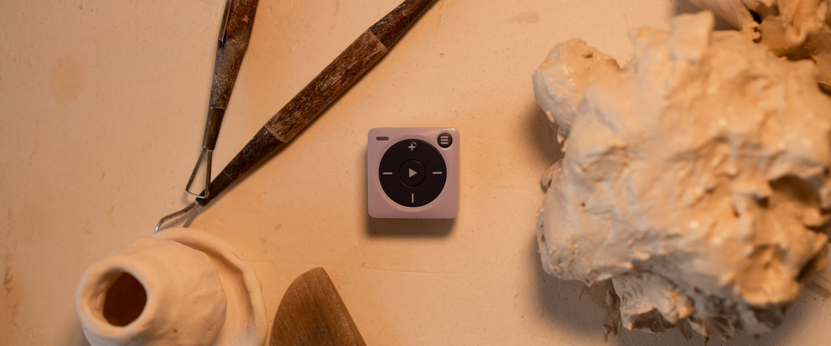 A photograph of a lavender Mighty 3 sitting on a table with tools used to shape pottery. A lump of clay, clay tools, and a mini hand made vase surround the device. 