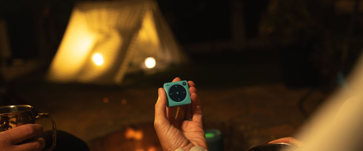 A night photo of a couple sitting in front of a campfire holding a blue Mighty 3 in hand. An occupied tent with a light on inside is in the background. The Mighty users' companion is clutching a copper Mule mug. 