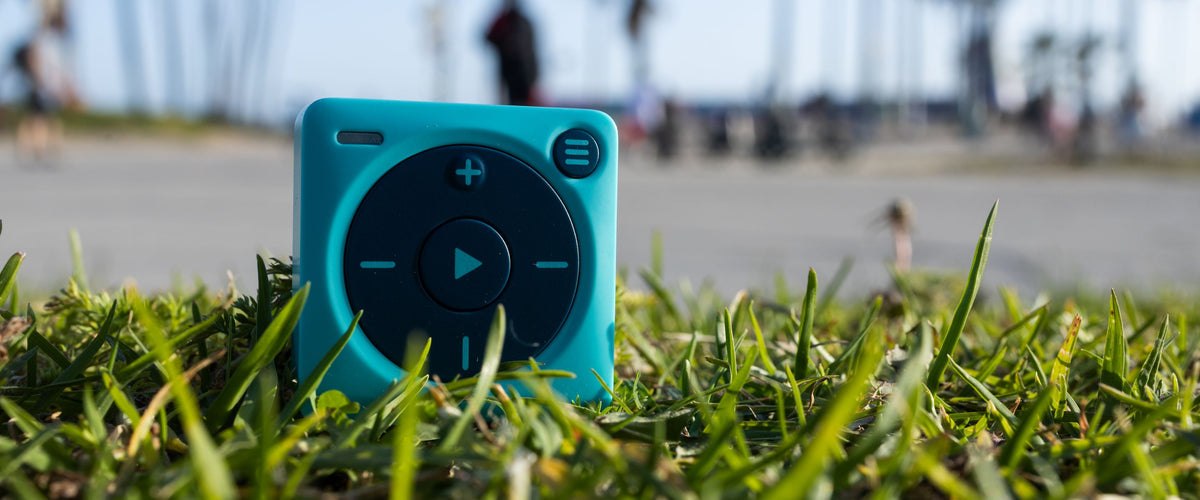 Close up photograph of blue Mighty 3 propped up on a patch of grass. A vignetted / blurred background of a beach walkway with people walking in the background. Macro-type image. 
